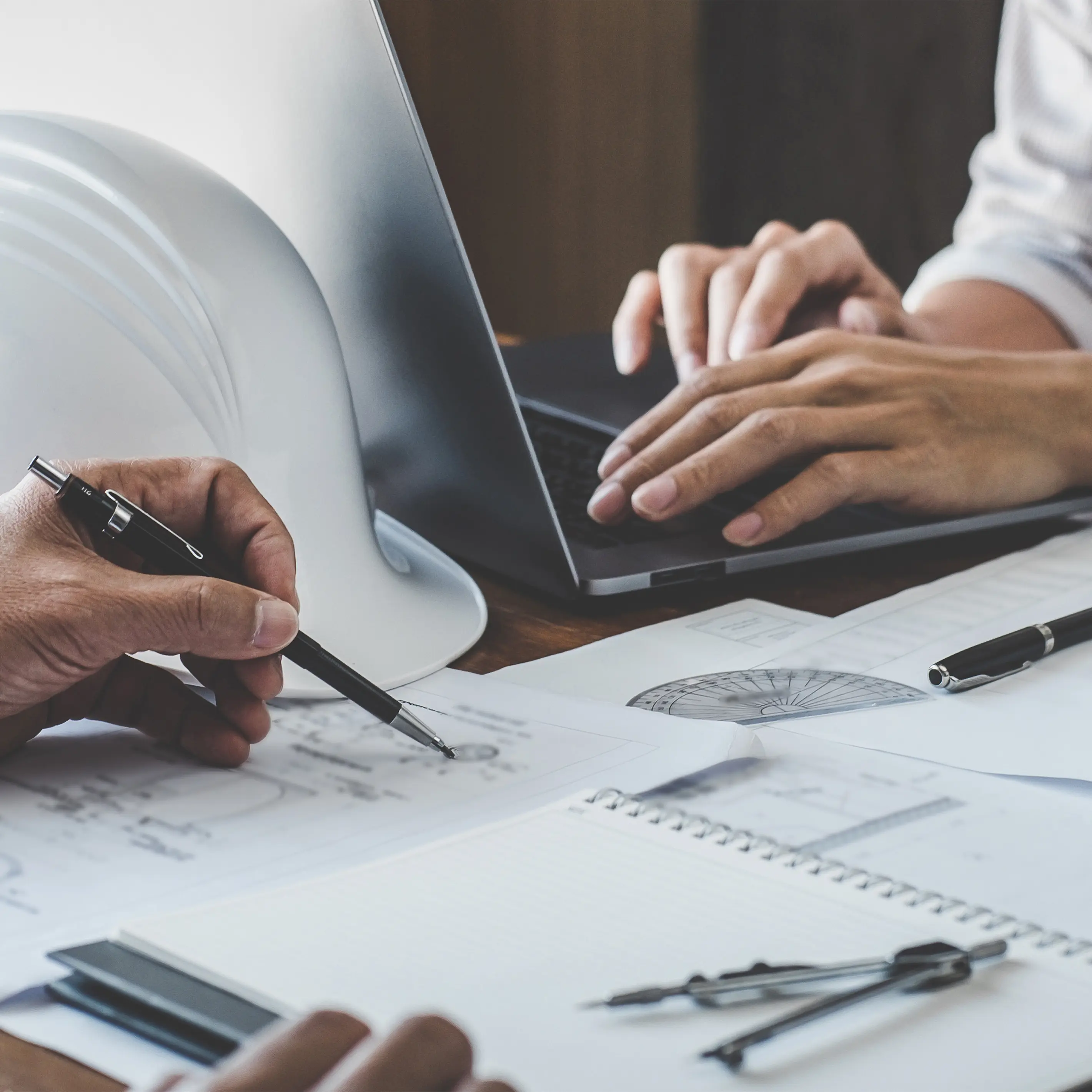 Escritorio de trabajo con una computadora, planos y un casco de construcción con manos de trabajadores
